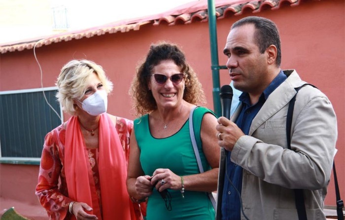 Matilde, vestita di verde e sorridente, posa accanto a una donna, in abito rosso che indossa una mascherina ffp2 e accanto a un uomo in giacca panna e camicia blu. L'uomo sta parlando al microfono