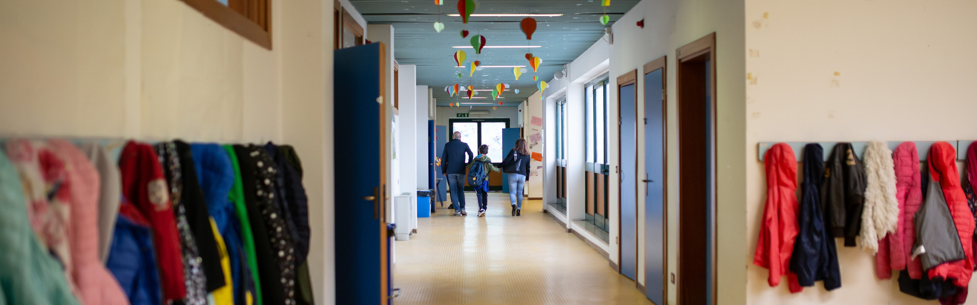 Francesco entra a scuola tenuto per mano da mamma e papà. Sono inquadrati di spalle mentre camminano lungo i corridoi