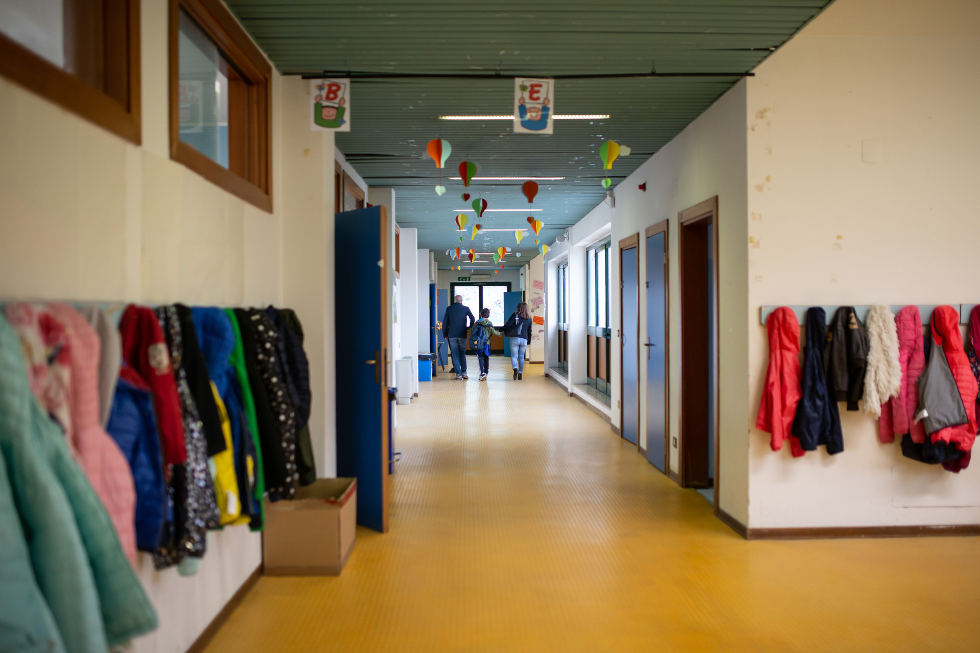 Francesco entra a scuola tenuto per mano da mamma e papà. Sono inquadrati di spalle mentre camminano lungo i corridoi 