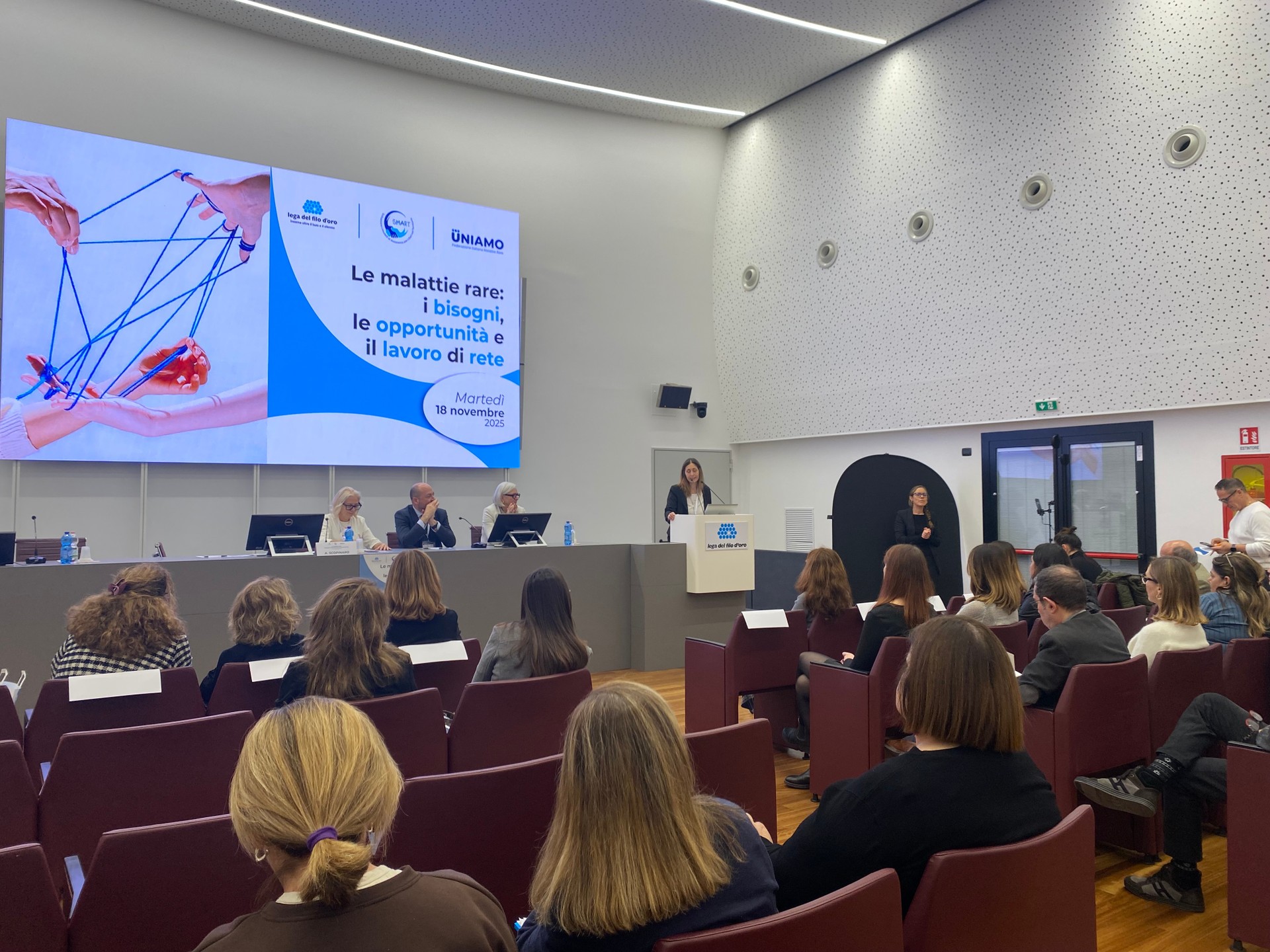 Un momento dell'evento sulle malattie rare tenutosi presso la sala polifunzionale della Lega del Filo d'Oro. I tre relatori seduti al tavolo e la platea ascoltano la dottoressa Santoro.