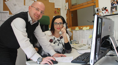 Andrea e Eleonora, due centralinisti della Lega del Filo d'Oro, sono fotografati mentre lavorano. Eleonora ha la cornetta del telefono appoggiata all'orecchio ed è seduta alla scrivania. Andrea sta lavorando al computer. Entrambi guardano verso la fotocamera.