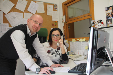 Andrea e Eleonora, due centralinisti della Lega del Filo d&#39;Oro, sono fotografati mentre lavorano. Eleonora ha la cornetta del telefono appoggiata all&#39;orecchio ed &#232; seduta alla scrivania. Andrea sta lavorando al computer. Entrambi guardano verso la fotocamera.
