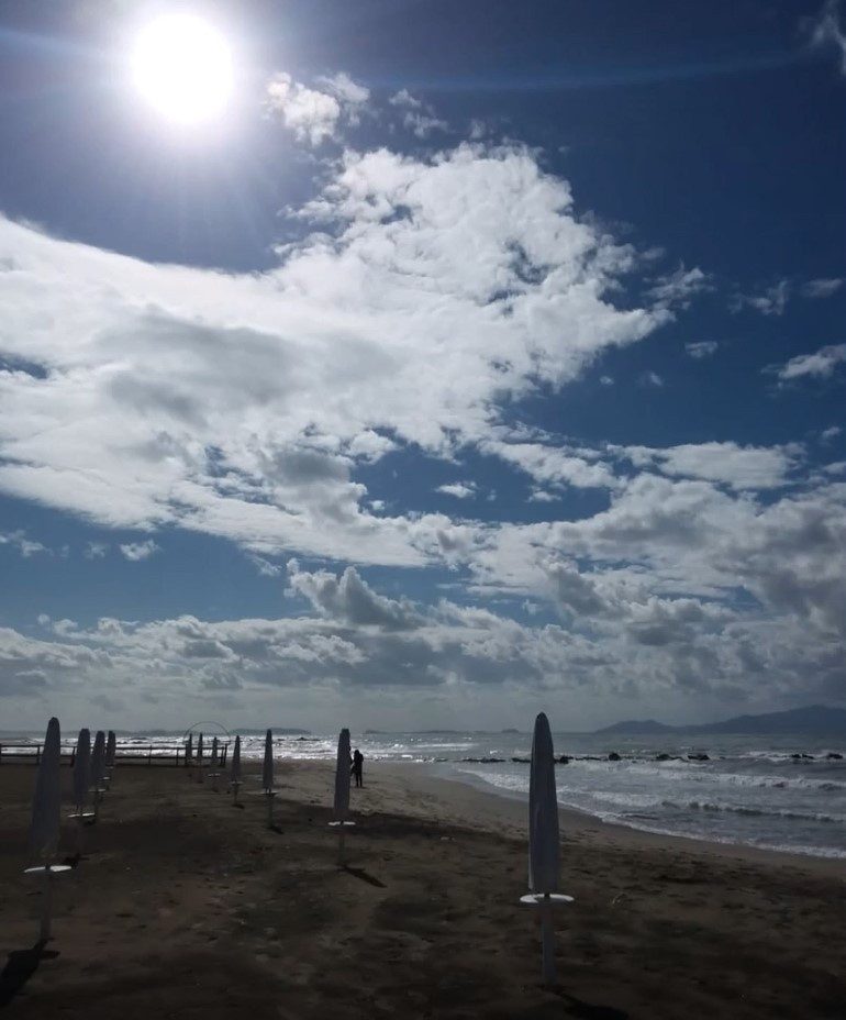 In primo piano c'è la spiaggia con gli ombrelloni chiusi. Il sole splende alto e illumina tutto. Il mare è agitato.