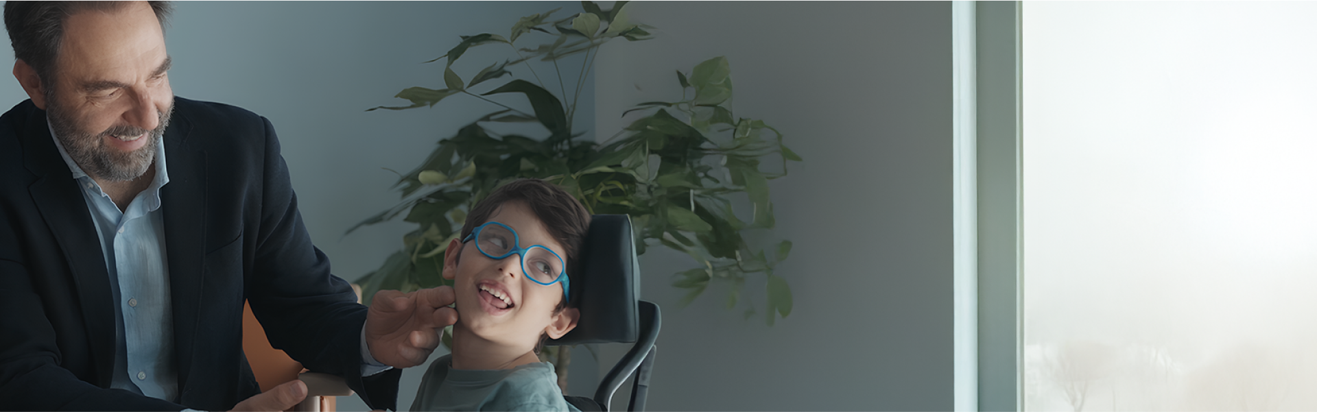 Foto di Neri Marcorè e di Nicola. Neri Marcorè è seduto, sta sorridendo e sta accarezzando la guancia di Nicola, un bambino di 6 anni in sedia a rotelle. Nicola ha problemi motori e visivi. Indossa un paio di occhiali azzurri. Sorride