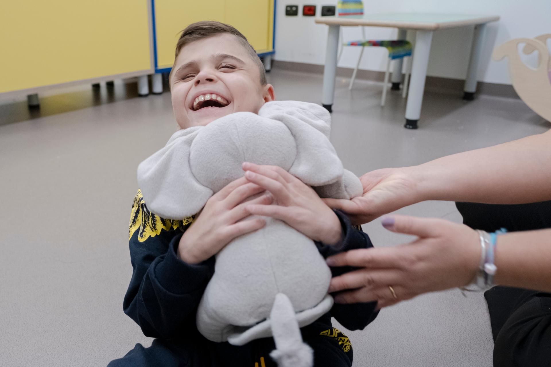 Primo piano di Matteo, bambino sordocieco e del suo elefanino di peluches. Lo sguardo di Matteo è frontale e sorride felice