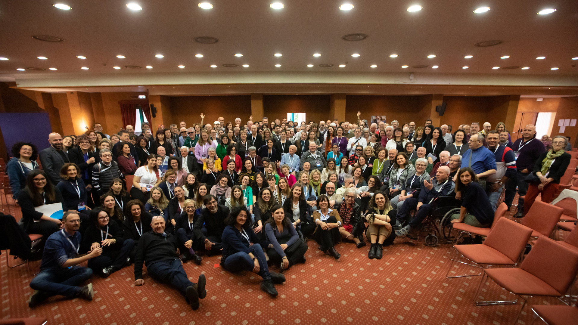 Foto di gruppo alla fine dell'Assemblea dei Sordociechi a TIvoli. In una grande sala con faretti sul soffitto e pavimento in moquette color aragosta con pois panna, c'è un gruppo di almeno un centinaio di persone che posa sorridente.