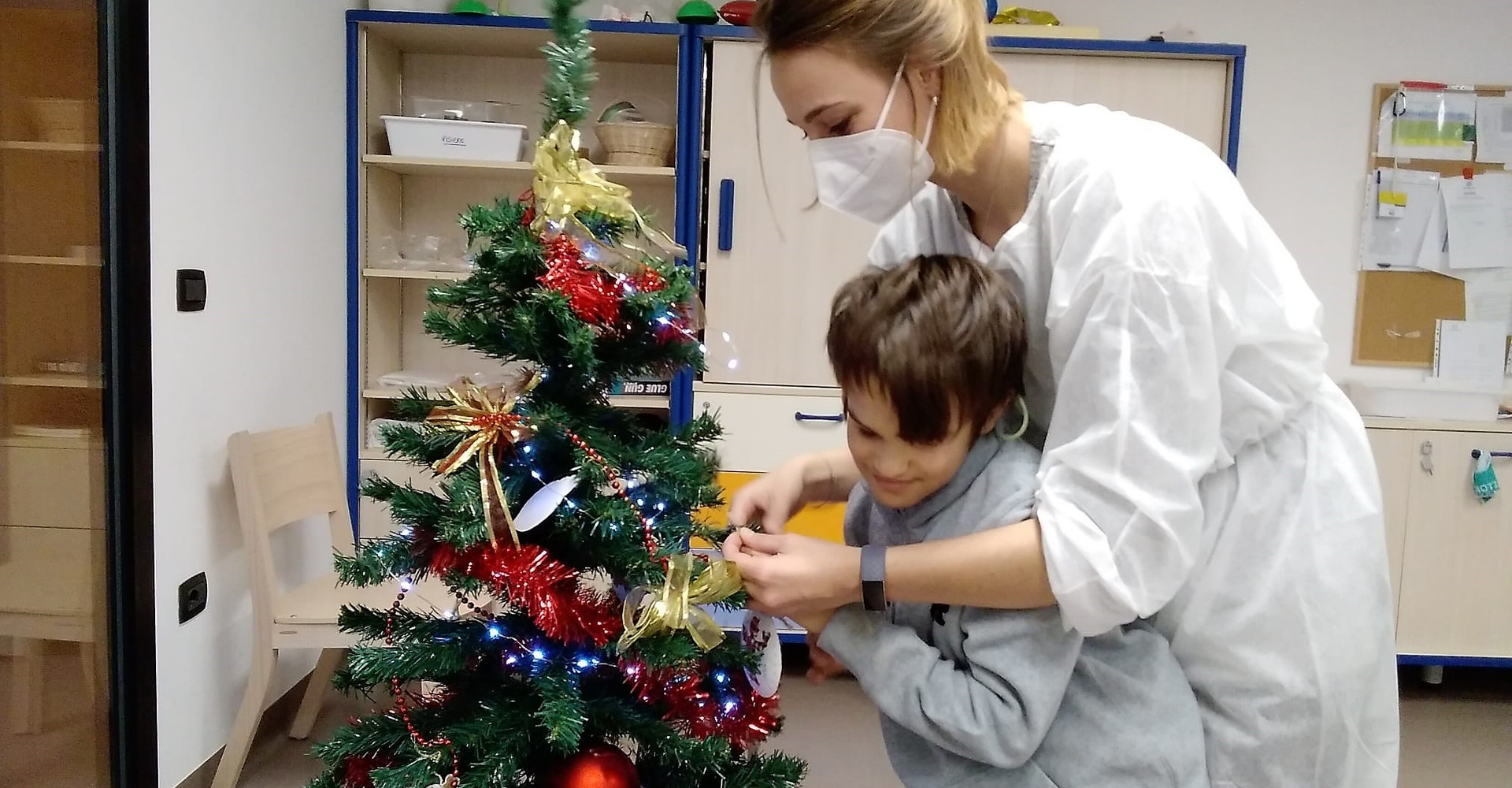 Un'operatrice e un bambino ospite del Centro Nazionale decorano un albero di Natale con palline e addobbi.