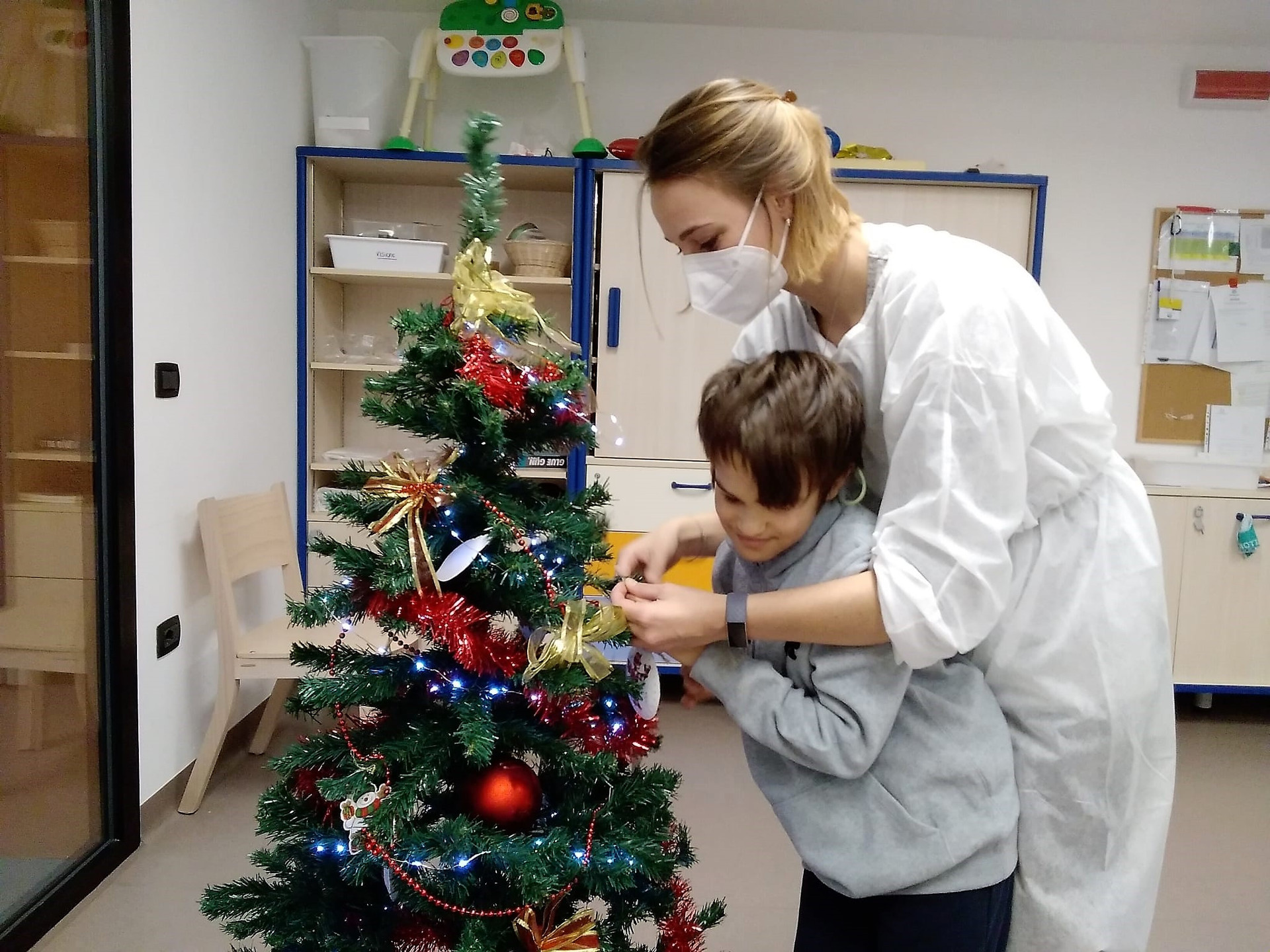 Un'operatrice e un bambino ospite del Centro Nazionale decorano un albero di Natale con palline e addobbi.
