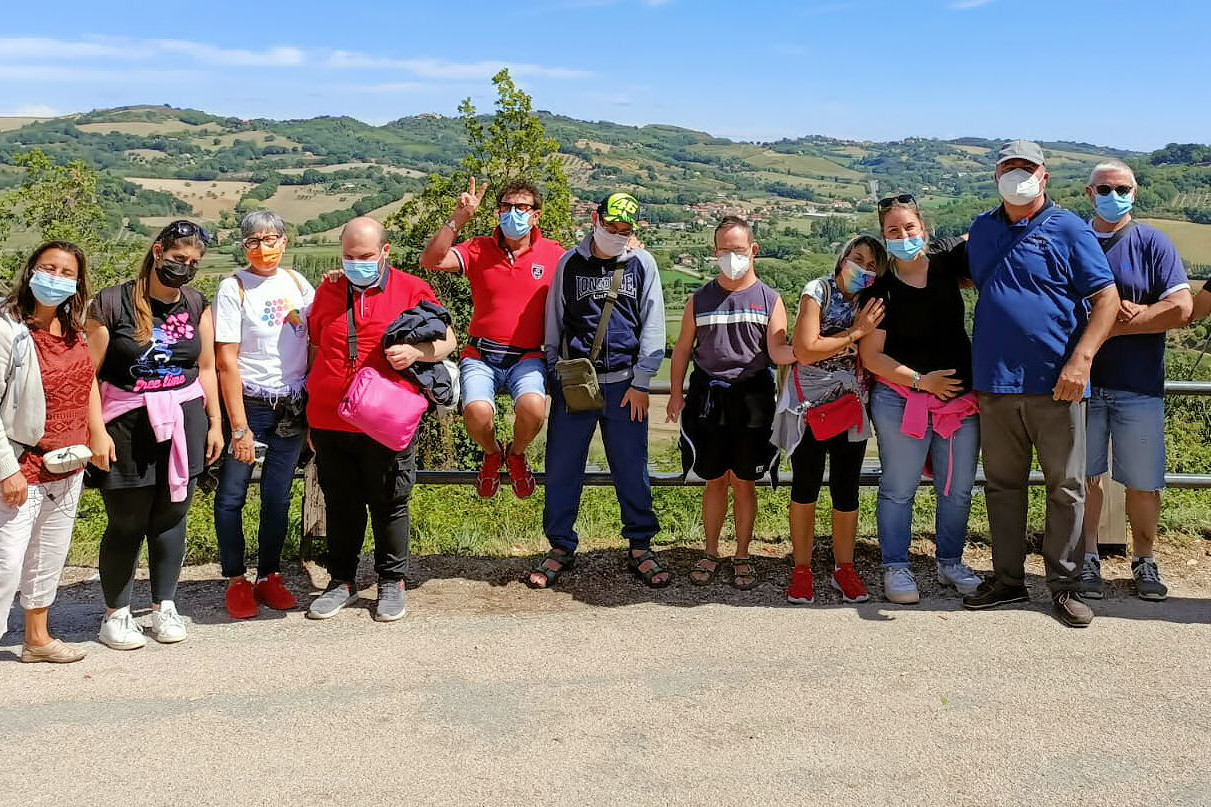 12 persone tra utenti, operatori e volontari posano in piedi per una foto di gruppo. Dietro di loro ci sono le colline.