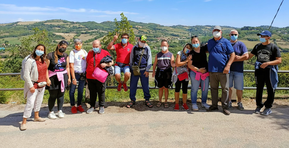12 persone tra utenti, operatori e volontari posano in piedi per una foto di gruppo. Dietro di loro ci sono le colline.