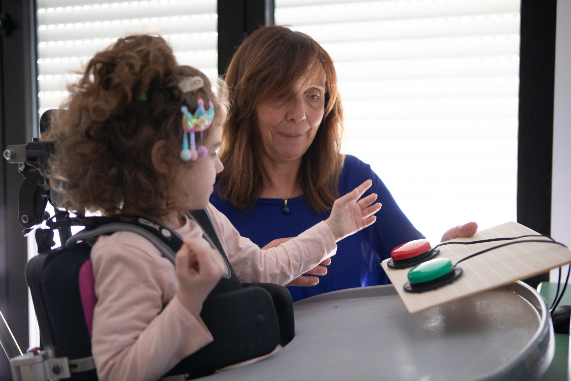 Melissa interagisce con due pulsanti switch, con l'aiuto e la supervisione di una donna adulta, forse un'educatrice o sua madre. Le due collaborano durante l'attività. Melissa è in primo piano, seduta su un seggiolone a cui è agganciata con cinture.