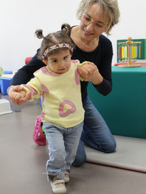 Sophie, bambina sordocieca con la sindrome di Charge, insieme alla sua educatrice.- L'educatrice è in ginocchio e sta aiutando Sophie a camminare