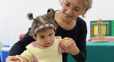 Sophie, bambina sordocieca con la sindrome di Charge, insieme alla sua educatrice.- L'educatrice è in ginocchio e sta aiutando Sophie a camminare