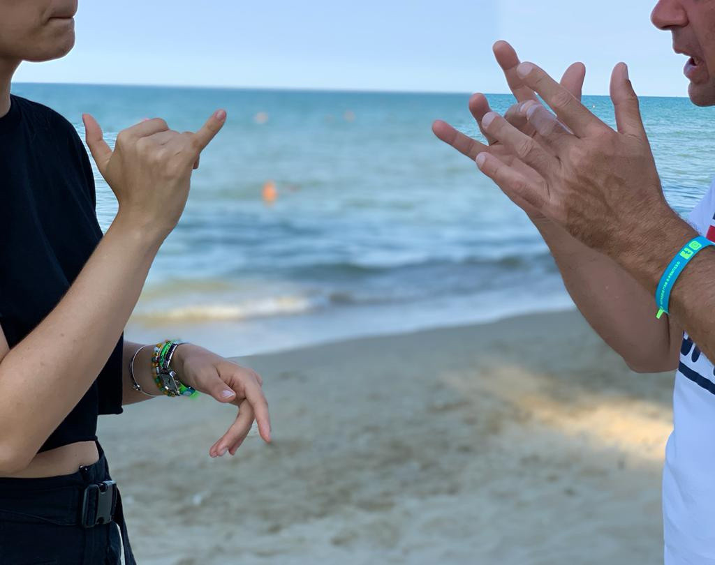 Primo piano di mani che comunicano in LIS (Lingua dei Segni Italiana) tra un’utente e una volontaria della Lega del Filo d’Oro. Sullo sfondo si intravedono la spiaggia e il mare. I volti non sono visibili, l’attenzione è sulle mani.