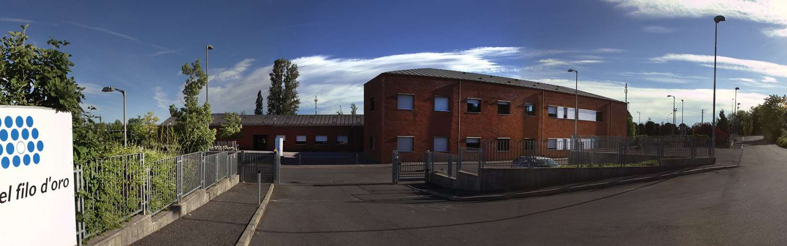 Foto a campo largo della Sede di Lesmo. A sinistra il cartello con il logo della Lega del Filo d'Oro e un cancello, mentre centralmente si vede l'edificio principale in mattoni rossi. La strada continua a si allontana sulla destra. Il sole splende nel cielo azzurro, con qualche nuvola.
