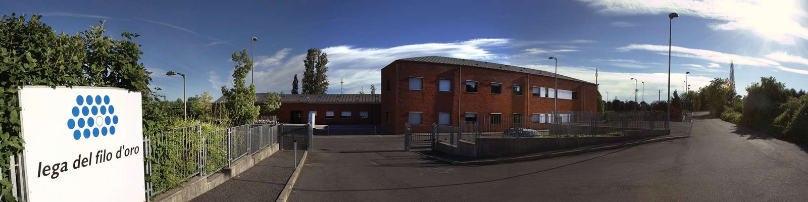 Foto a campo largo della Sede di Lesmo. A sinistra il cartello con il logo della Lega del Filo d'Oro e un cancello, mentre centralmente si vede l'edificio principale in mattoni rossi. La strada continua a si allontana sulla destra. Il sole splende nel cielo azzurro, con qualche nuvola.