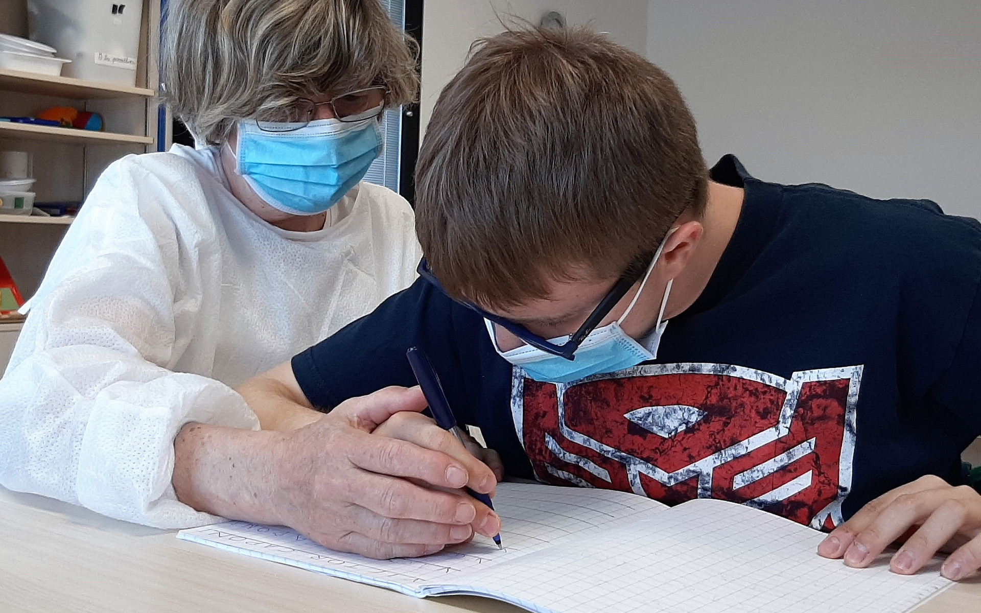 Victor scrive su un quaderno con la sua educatrice seduta accanto a lui al banco. L'educatrice guida la mano di Victor con la propria. Entrambi indossano le mascherine chirurgiche.