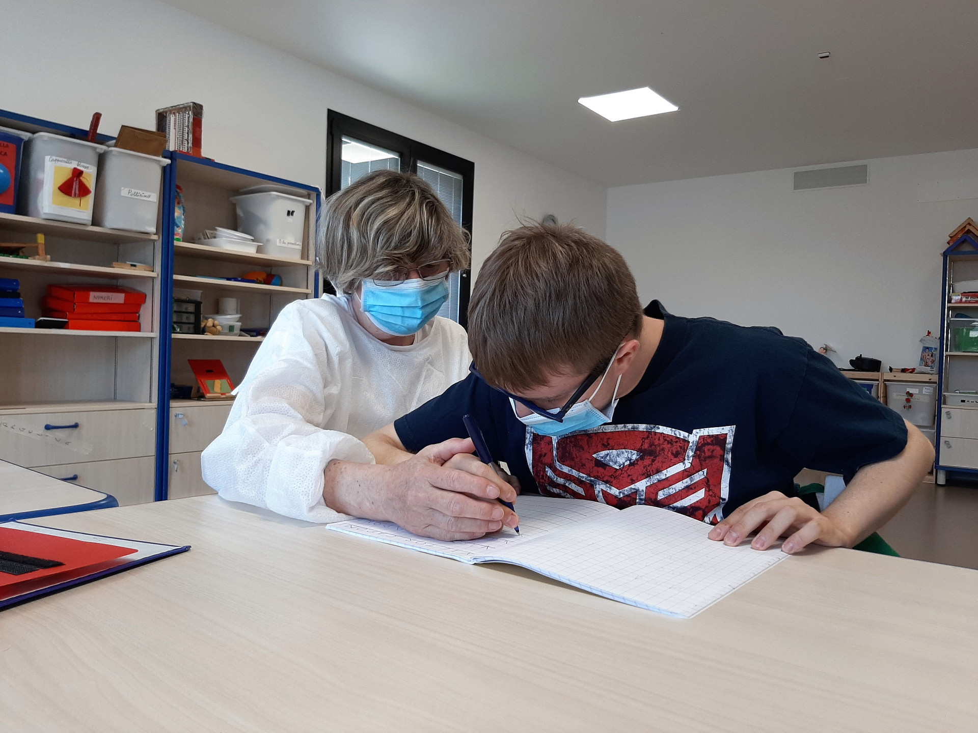 Victor scrive su un quaderno con la sua educatrice seduta accanto a lui al banco. L'educatrice guida la mano di Victor con la propria. Entrambi indossano le mascherine chirurgiche.