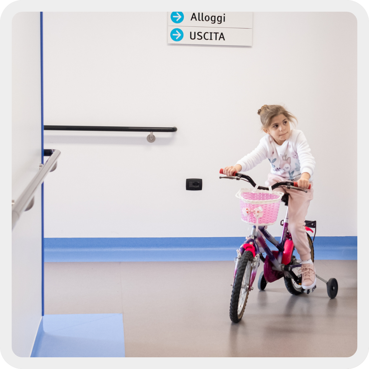 Sveva, bambina con Sidnrome di Charge con maglia bianca e pantaloni rosa, gira per i corridoio del Centro Nazionale con la sua bicicletta fucsia.