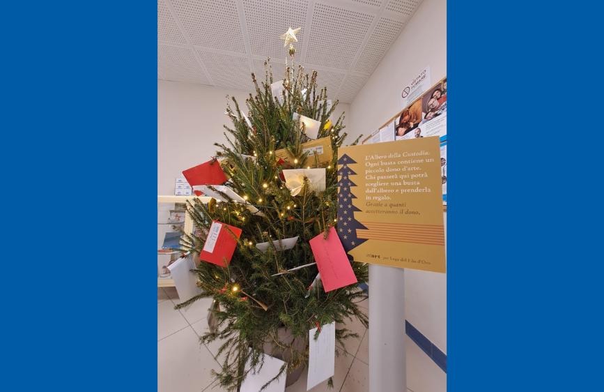 Albero di Natale decorato con luci e buste colorate contenenti messaggi dono. Accanto, un cartello spiega l’iniziativa solidale ‘L’Albero della Custodia’ a favore della Lega del Filo d’Oro.