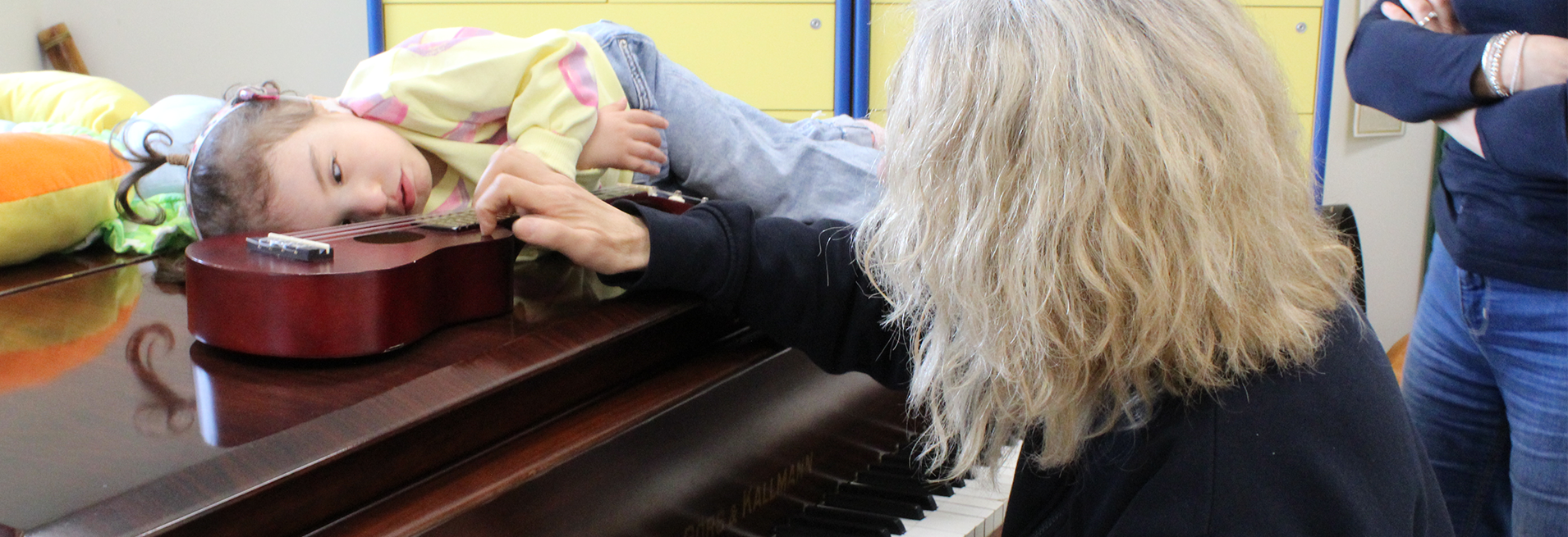 Seduta di musicoterapia. Sophie, una bambina sordocieca di circa 3 anni è distesa sul pianoforte. Di fronte a lei, c'è una piccola chitarra. Fiammetta, la musicoterapista, pizzica le corde della chiatarra. Fimmetta è seduta al pianoforte. E' di spalla 