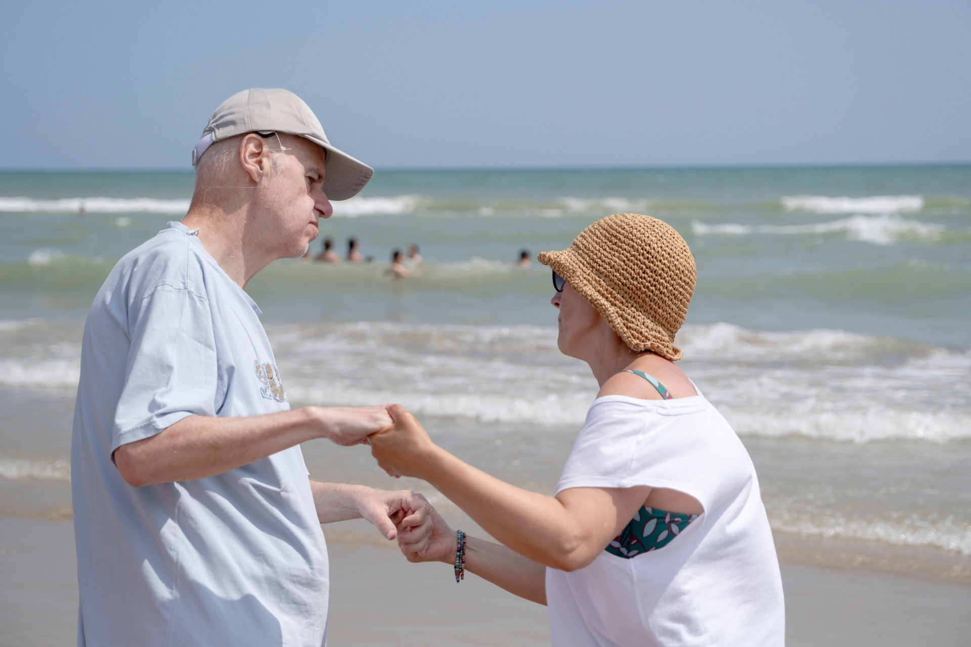 In riva al mare, una volontaria tiene per mano un uomo mentre ballano insieme, circondati dalla brezza estiva e dalla gioia del soggiorno: un momento di leggerezza, connessione e libertà condivisa.