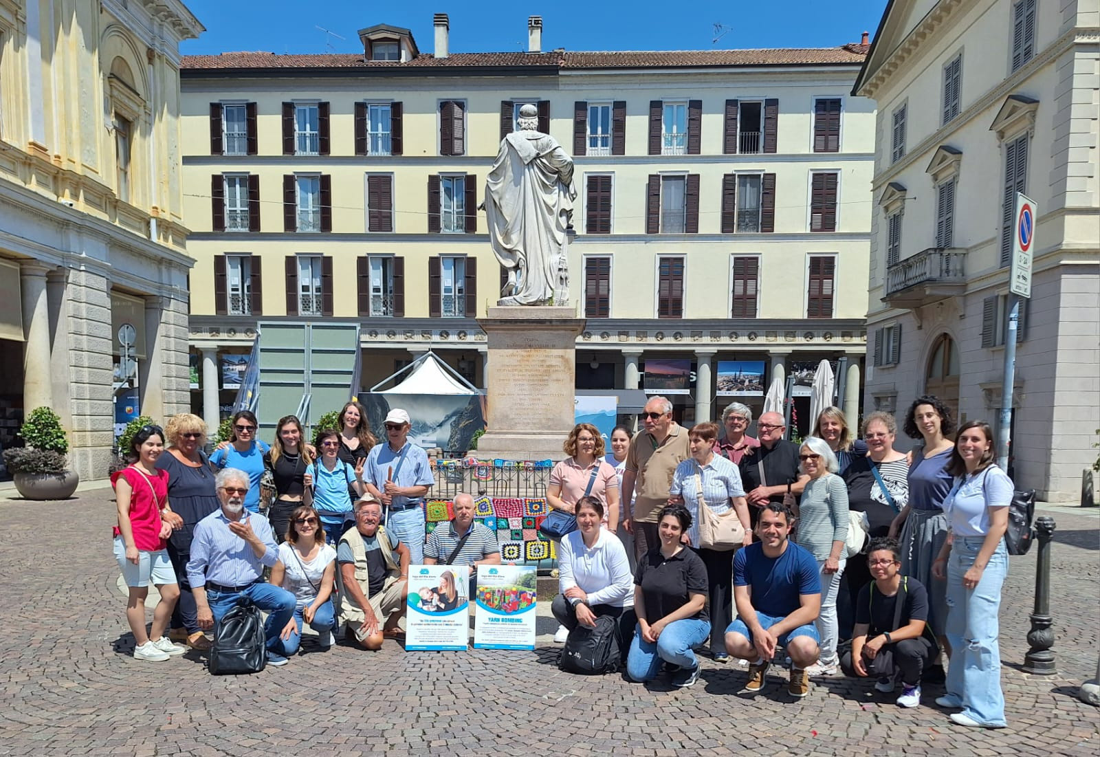 Un gruppo di volontari posa con entusiasmo in una piazza decorata per lo Yarn Bombing. Alle loro spalle si erge una statua.