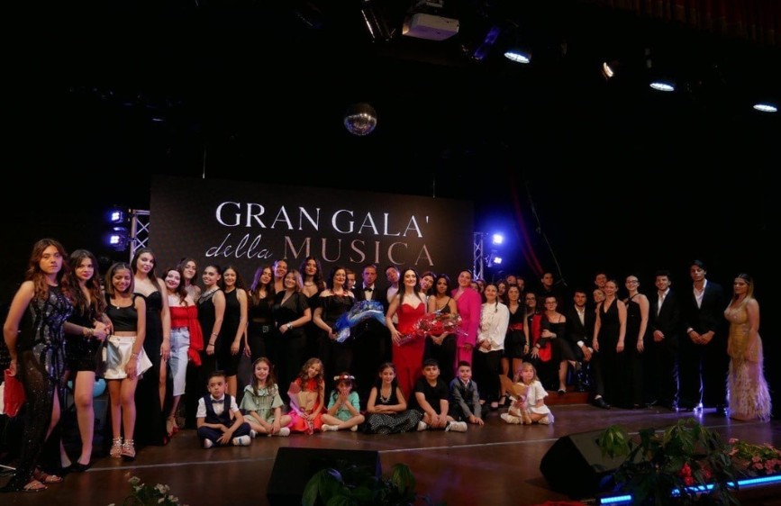 L'immagine mostra un numeroso gruppo di persone, tra cui diversi bambini seduti in prima fila, sul palco di un evento serale intitolato "Gran Galà della Musica" tenutosi il 4 luglio 2023. Sullo sfondo, un grande schermo nero con la scritta "GRAN GALA' della MUSICA" in bianco è illuminato, e una palla stroboscopica pende dal soffitto, suggerendo un'atmosfera festosa e celebrativa. La maggior parte degli adulti indossa abiti eleganti scuri, mentre alcuni si distinguono con abiti rossi o chiari.
