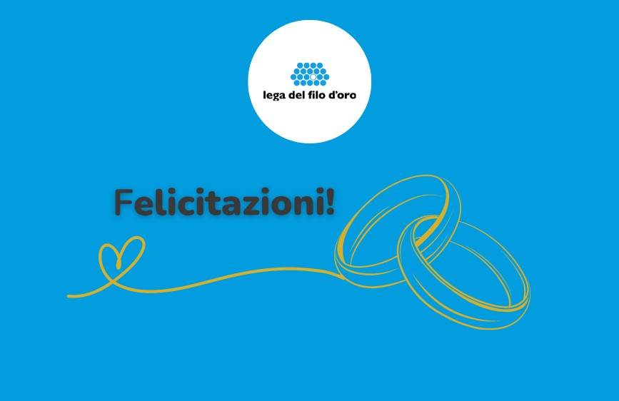 L'immagine mostra un biglietto di auguri su sfondo blu brillante. In alto al centro c'è il logo della "Lega del Filo d'Oro". Al centro, in grandi lettere marroni, si legge "Felicitazioni!", accompagnato da un piccolo cuore stilizzato dorato che si unisce a un paio di fedi nuziali dorate intrecciate, posizionate in basso a destra.