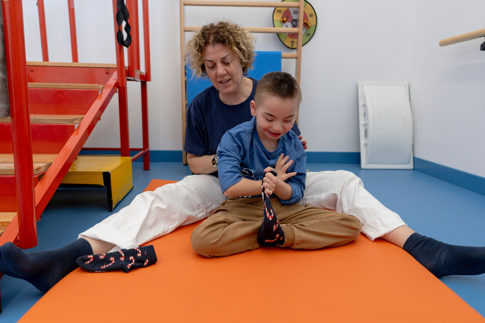Raffaella ed Edoardo sono insieme durante la fisioterapia. Edoardo sta cercando di togliersi un calzino da solo, con l'aiuto di Raffaella che lo sostiene mantenendo la schiena dritta, permettendo a Edoardo di concentrare tutte le sue forze per togliersi il calzino.
