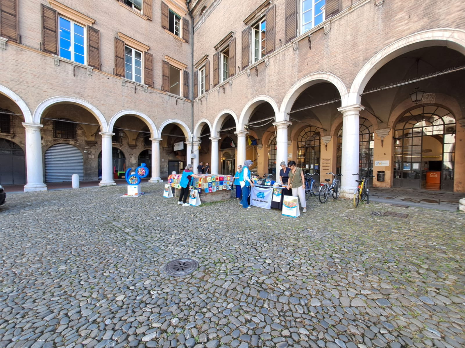 Panoramica di un angolo di piazza allestito per lo Yarn Bombing. Alcune persone stanno montando lo stand della Lega del Filo d’Oro, tra decorazioni colorate e materiali tessili.