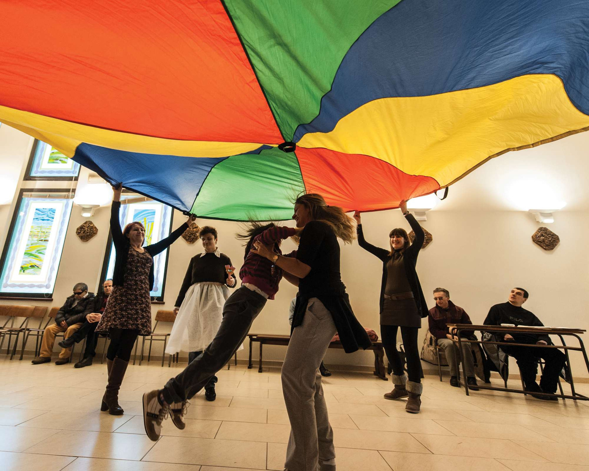 In un'ampia sala, ci sono tante persone. Sullo sfondo, seduti sulle sedie, ci sono due utenti adulti e due uomini. In piedi, davanti alla fotocamera, ci sono invece quattro donne e una ragazza, che festeggiano saltando e muovendosi. Due di loro reggono un telo che si estende sopra le teste di tutte le donne in piedi e che è colorato a spicchi gialli, rossi, verdi e blu.