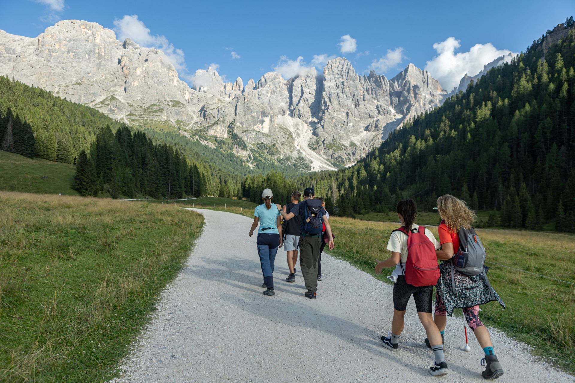 Alcuni partecipanti del soggiorno estivo in montagna organizzato dalla Lega del Filo d’Oro camminano lungo una strada sterrata immersa nel verde. Intorno si estendono prati e boschi, mentre sullo sfondo si stagliano alte montagne rocciose sotto un cielo azzurro.