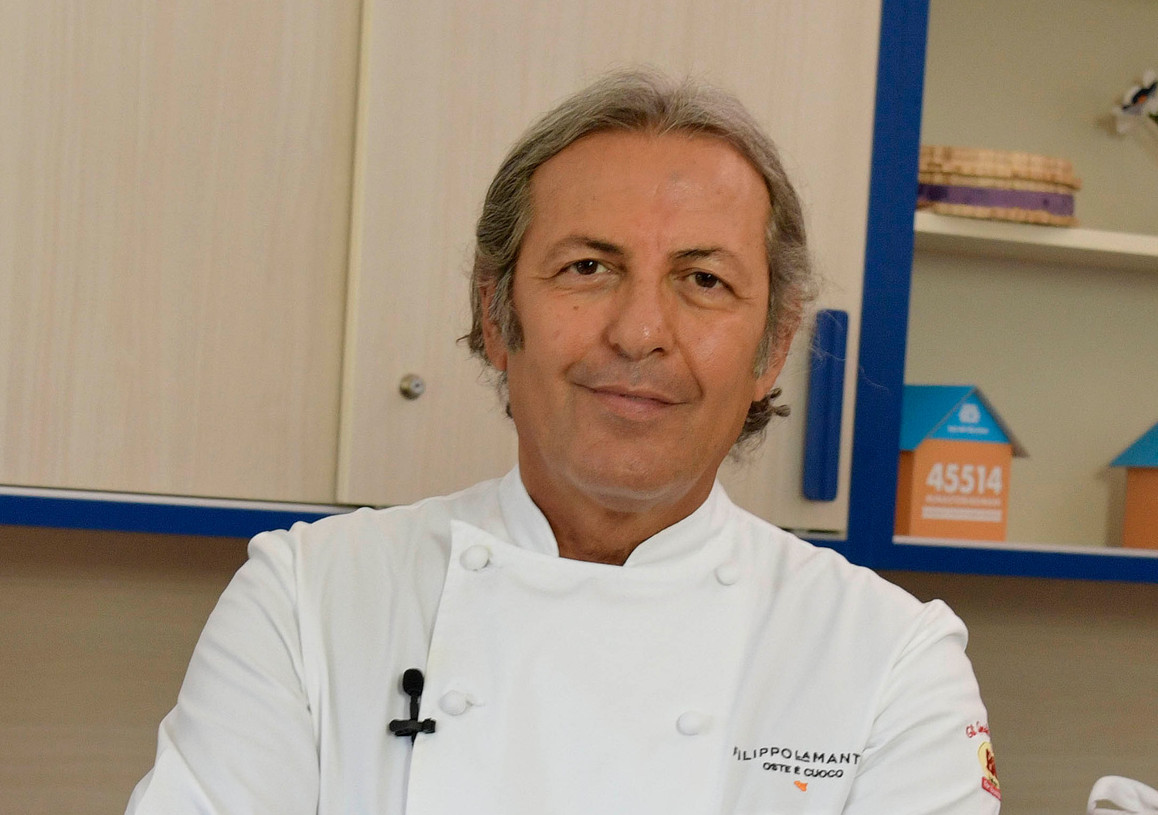 Lo chef La Mantia è nella cucina didattica del Centro di Lesmo insieme a Francesca, Maria Assunta e Giovanni, utenti adulti con cui sta per cucinare insieme, sul piano di lavoro c'è la casetta segnalino della campagna #uncontattochevale con il numero per donare via SMS solidale