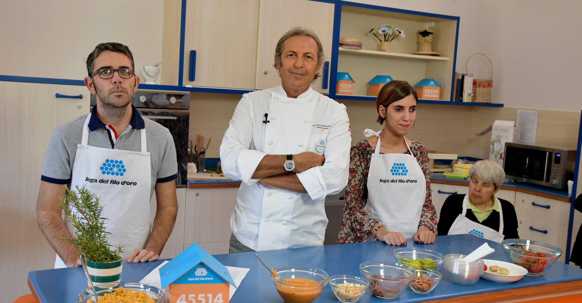 Lo chef La Mantia è nella cucina didattica del Centro di Lesmo insieme a Francesca, Maria Assunta e Giovanni, utenti adulti con cui sta per cucinare insieme, sul piano di lavoro c'è la casetta segnalino della campagna #uncontattochevale con il numero per donare via SMS solidale