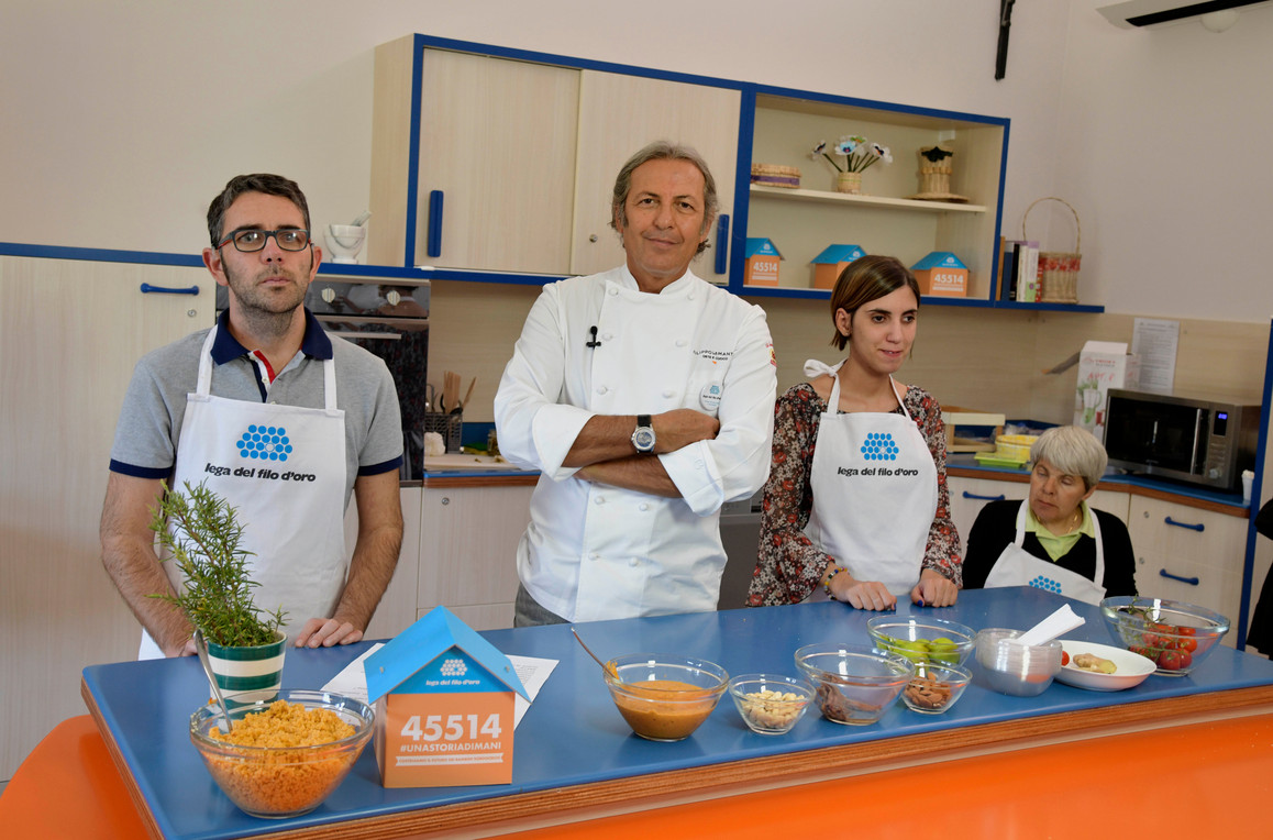 Lo chef La Mantia è nella cucina didattica del Centro di Lesmo insieme a Francesca, Maria Assunta e Giovanni, utenti adulti con cui sta per cucinare insieme, sul piano di lavoro c'è la casetta segnalino della campagna #uncontattochevale con il numero per donare via SMS solidale