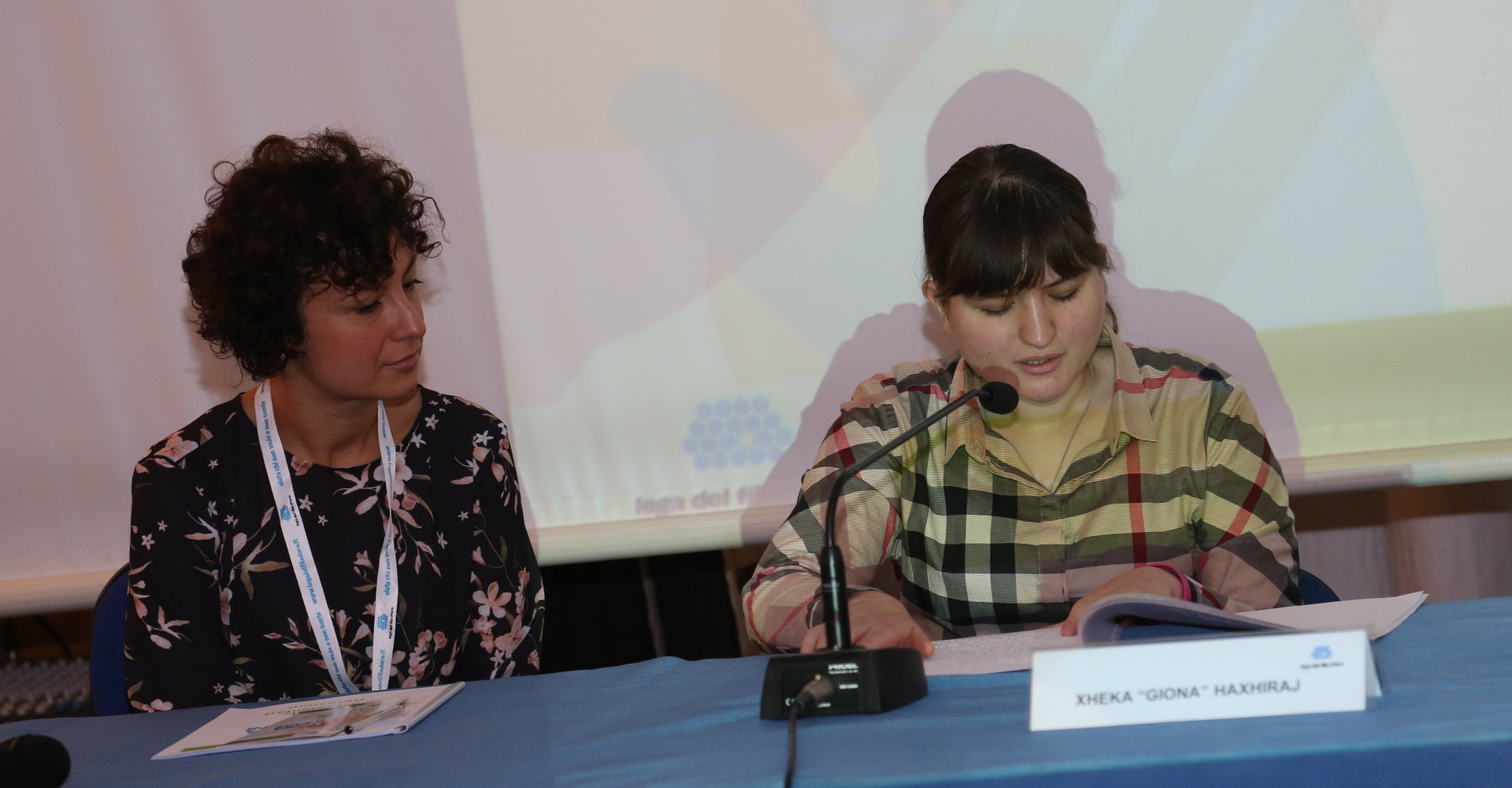 Giona, ragazza sordocieca, parla durante un evento della Lega del Filo d'Oro. E' seduta a un tavolo e davanti a lei ha un microfono e un cartello con scritto il suo nome. Una dipendente della "Lega" è seduta accanto a lei e la ascolta.