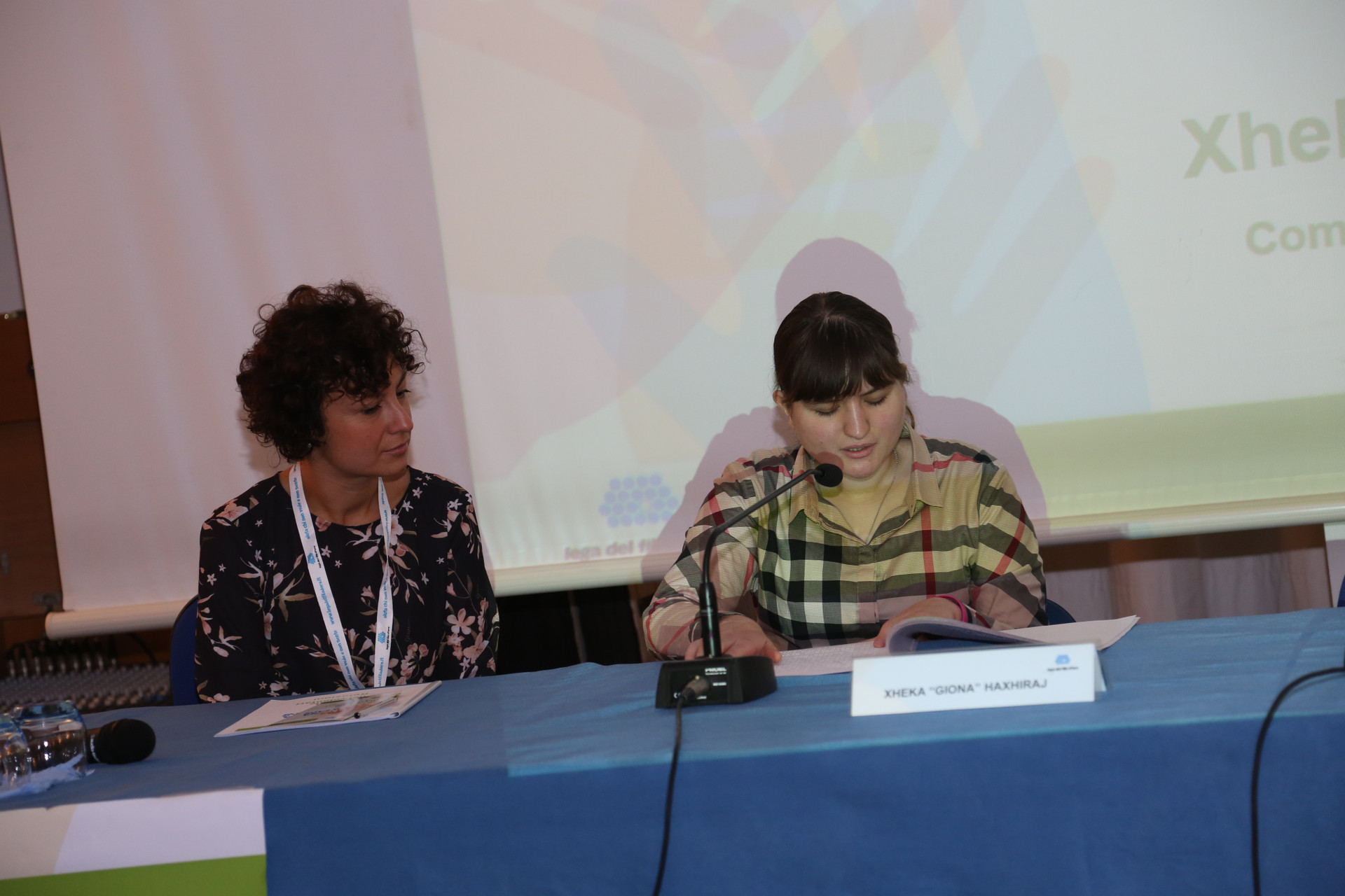 Giona, ragazza sordocieca, parla durante un evento della Lega del Filo d'Oro. E' seduta a un tavolo e davanti a lei ha un microfono e un cartello con scritto il suo nome. Una dipendente della "Lega" è seduta accanto a lei e la ascolta.