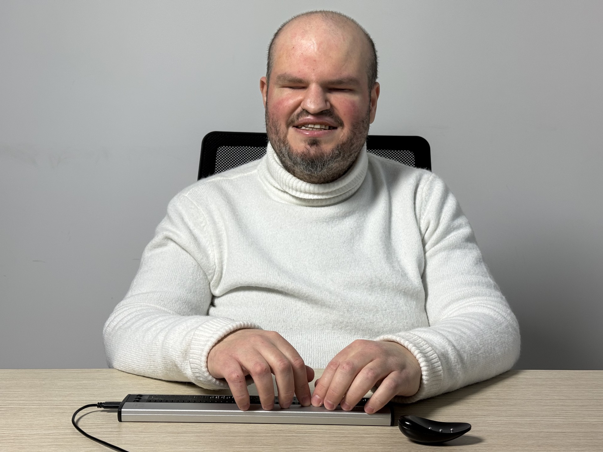 Francesco è seduto alla scrivania e digita su una barra braille. Indossa un maglione bianco a collo alto. 