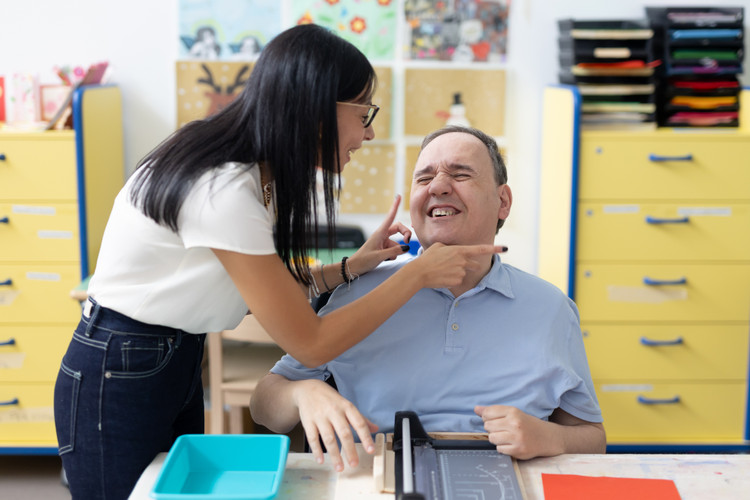 Alberto sorride accanto a un’operatrice che gli sfiora delicatamente il viso, creando un momento di dolcezza e vicinanza.