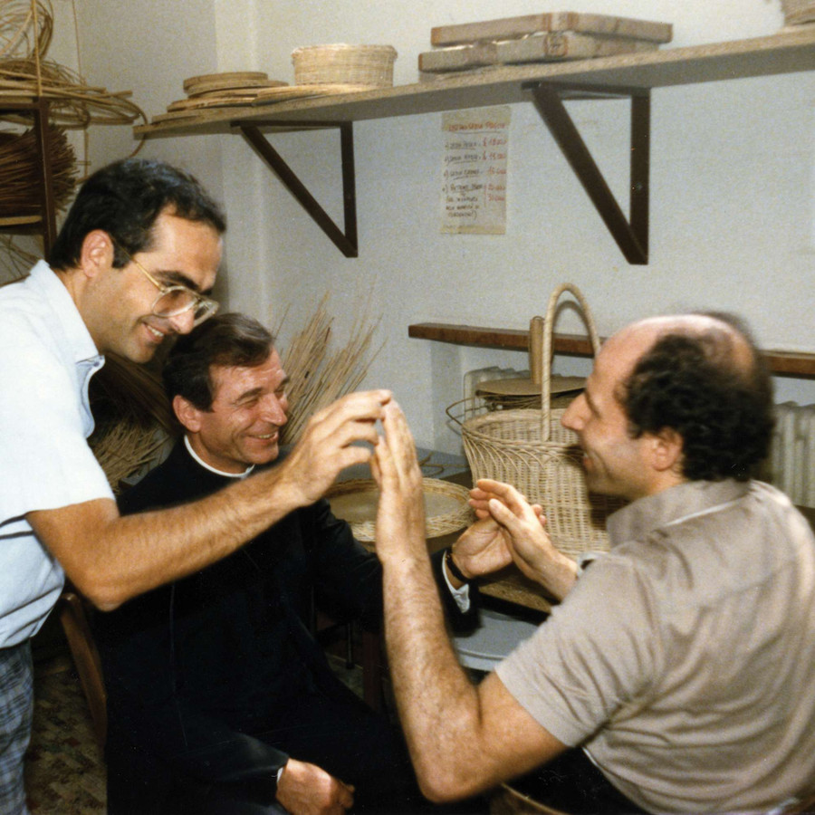 Un utente sordocieco incontra il Monsignor Francesco Casalini e un altro uomo, che qui saluta con tocco di mano. Tutti e tre sono sorridenti.