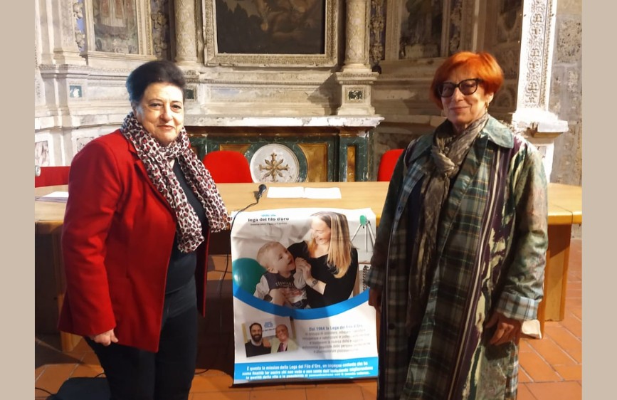 Due donne in piedi ai lati di un tavolo in una sala storica con pareti affrescate. Al centro, un grande manifesto con il logo della Lega del Filo d’Oro e la foto di una donna con un bambino sorridente.