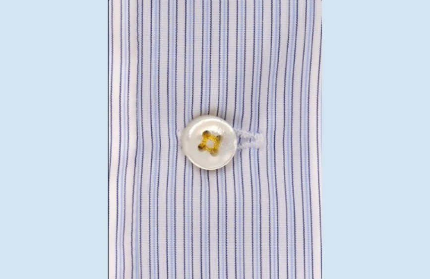 Primo piano di un bottone bianco cucito con filo giallo su una camicia a righe sottili verticali blu e bianche.