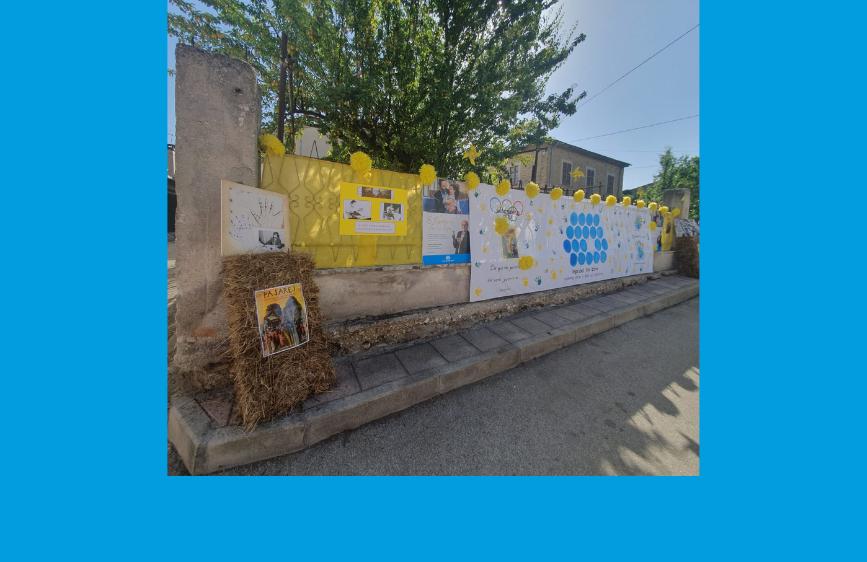 L'immagine mostra una recinzione di un muretto esterno, decorata con striscioni e poster promozionali delle "Quartieriadi 2023 San Benedetto dei Marsi". I materiali esposti, principalmente nei colori bianco e giallo, includono scritte e disegni che suggeriscono un evento comunitario e sportivo, con il logo della Lega del Filo d'Oro visibile su alcuni. Le decorazioni sono adornate con fiocchi gialli e in un angolo è posizionata una balla di fieno, aggiungendo un tocco rustico all'allestimento.