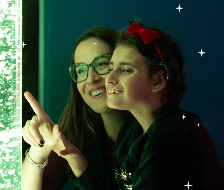 Aurora, bambina con disabilità uditive, sorride con la sua educatrice mentre indicano un fascio di luce nella stanza di stimolazione visiva. Momento di educazione inclusiva e apprendimento sensoriale.