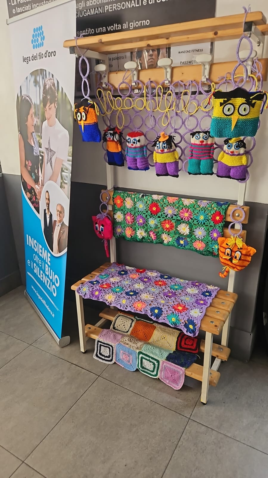 Una panchina da spogliatoio, all’interno di una palestra a Napoli, è decorata con filati colorati e pupazzi realizzati a maglia. Accanto si trova lo stand della Lega del Filo d’Oro, con il manifesto che riporta il claim “Insieme oltre il buio e il silenzio”.