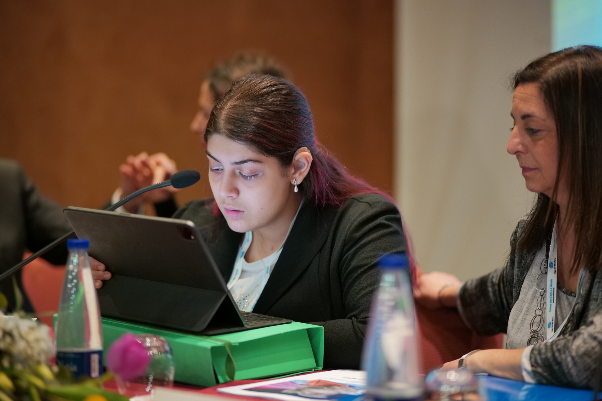 Lucia, giovane sordocieca, mentre espone il suo intervento durante una delle Conferenza delle Persone Sordocieche organizzata dalla Lega del Filo d'Oro. Lucia è affiancata dall'educatrice Cinzia che la sta ascoltando. Sullo sfondo si vedono una persona sordocieca che segue l'intervento di Lucia coadiuvata dall'interprete di LIS tattile.
