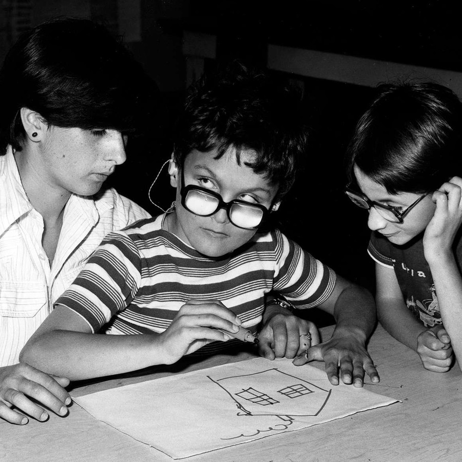 Foto scattata negli anni '70 e quindi in bianco e nero. Un bambino ospite dell'Istituto di Riabilitazione di Osimo sta disegnando una casa con un pennarello, seduto sulle gambe di un'operatrice. Un altro bambino guarda incuriosito.