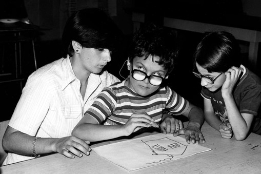 Foto scattata negli anni '70 e quindi in bianco e nero. Un bambino ospite dell'Istituto di Riabilitazione di Osimo sta disegnando una casa con un pennarello, seduto sulle gambe di un'operatrice. Un altro bambino guarda incuriosito.
