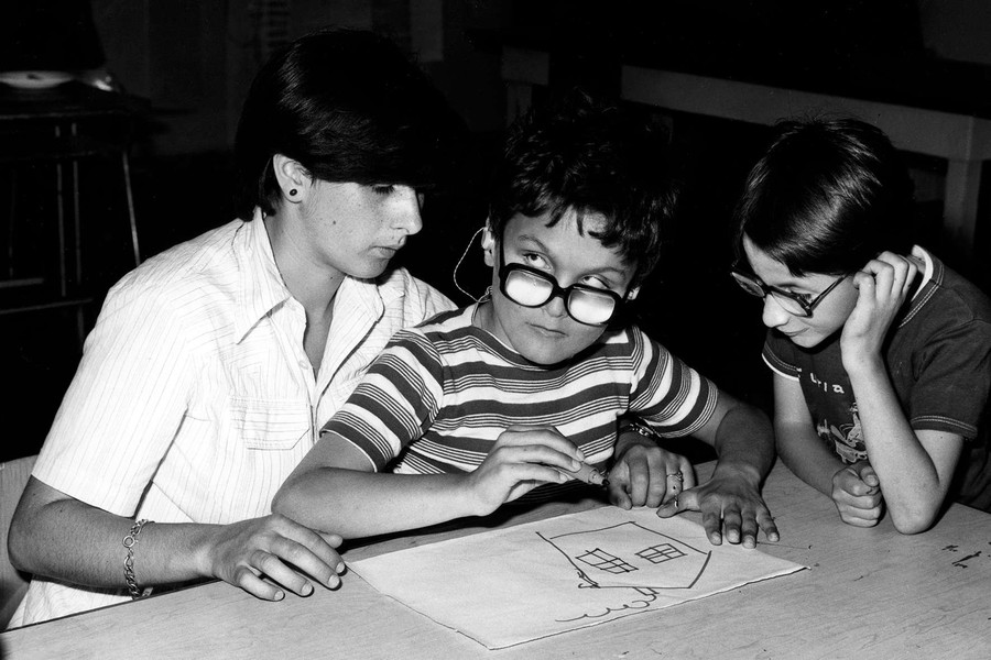 Foto scattata negli anni '70 e quindi in bianco e nero. Un bambino ospite dell'Istituto di Riabilitazione di Osimo sta disegnando una casa con un pennarello, seduto sulle gambe di un'operatrice. Un altro bambino guarda incuriosito.